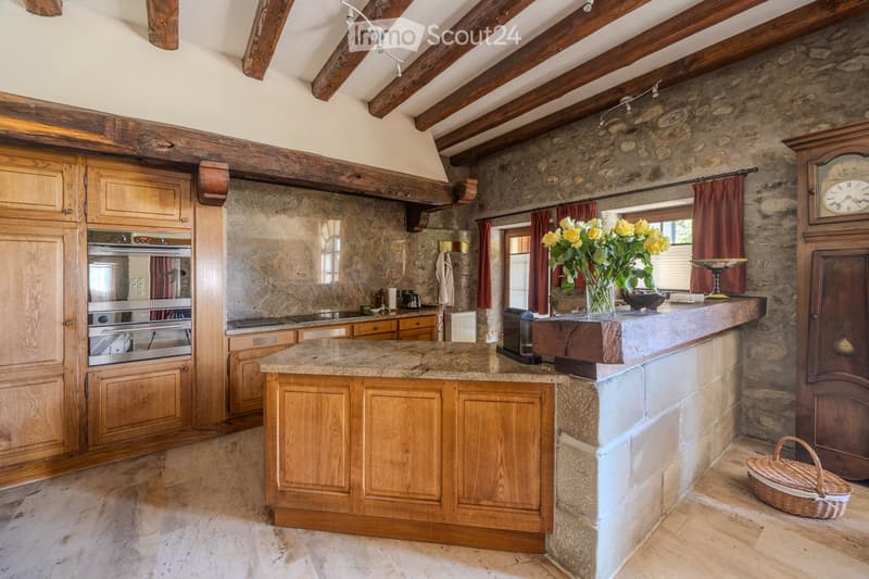 A rustic kitchen with exposed wooden beams, stone walls, and a central island with wooden cabinets and a granite countertop. Features include an oven, a vase with yellow flowers, a large clock, and a wicker basket on the floor.