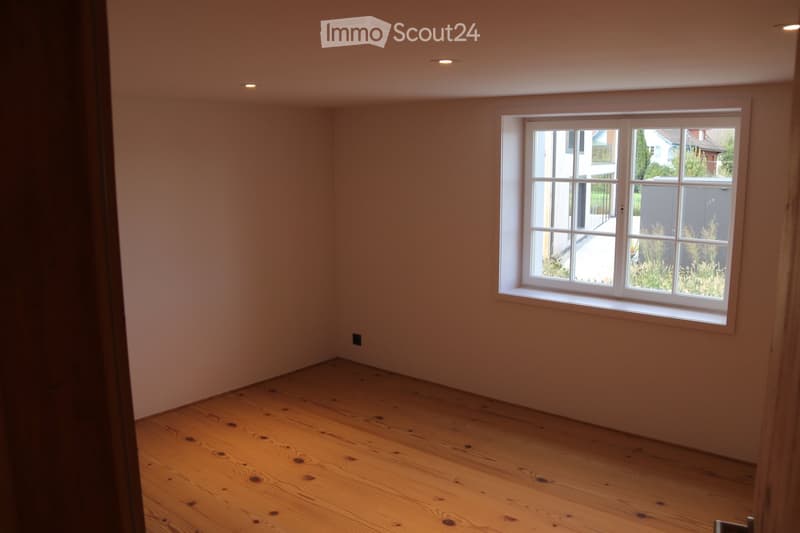 Empty room with wooden flooring and a large window showing a view of the outside. The walls are painted in a light neutral color, and the room has recessed lighting.