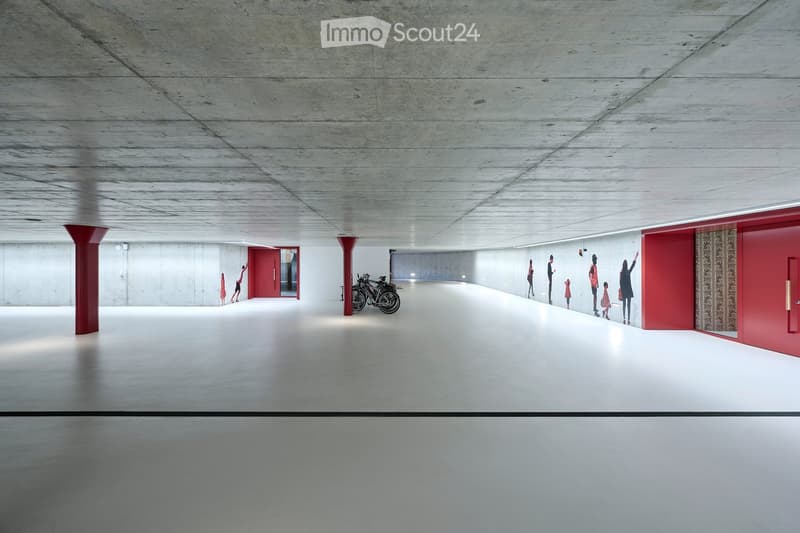 A spacious, modern corridor with a smooth concrete floor and ceiling, red accent walls, and a few people walking. The area is well-lit with recessed lighting and features red structural columns and doors.