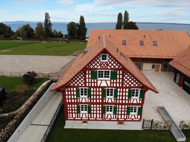 Traditional half-timbered house with red and white exterior, green shutters, and a red-tiled roof, situated near a body of water with a landscaped garden and open fields in the background.