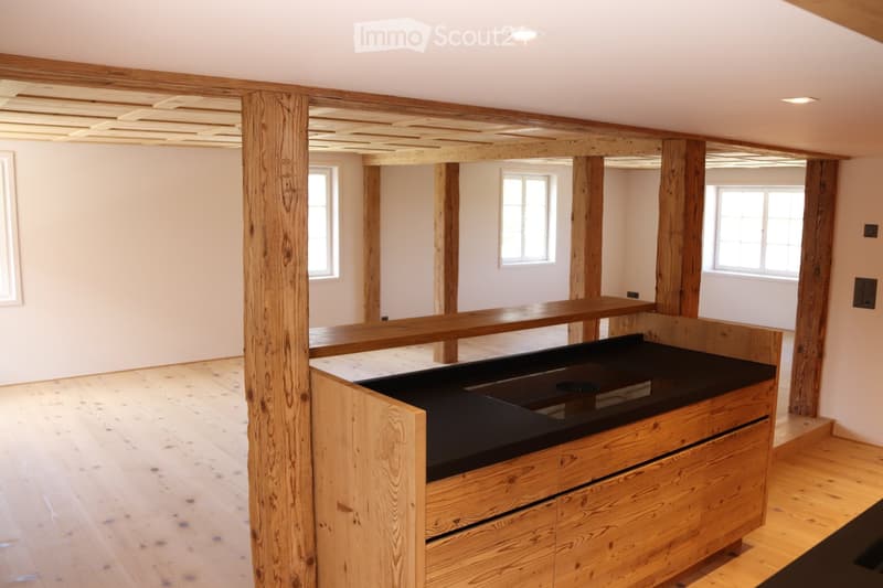 An unfurnished room with exposed wooden beams and a wooden floor, featuring a black countertop and wooden cabinetry, with large windows allowing natural light to enter.