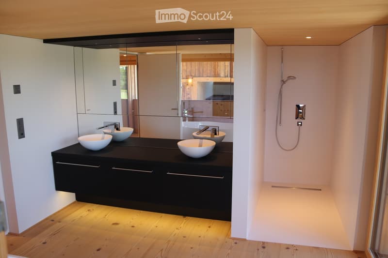 Modern bathroom with a sleek black vanity, four white sinks, and a large mirror. The shower area is visible on the right with a modern showerhead and controls.