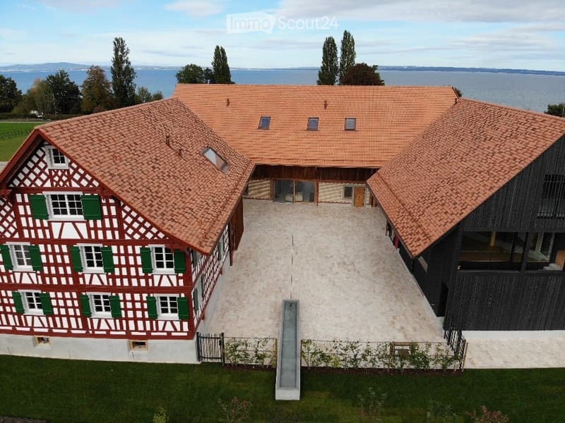 A large, modern building complex with a traditional red-tiled roof and green shutters, surrounded by a paved courtyard and greenery, with a view of a lake in the background.