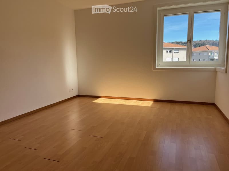 Empty room with light wooden flooring and white walls, featuring a large window with a view of neighboring buildings and a clear blue sky.