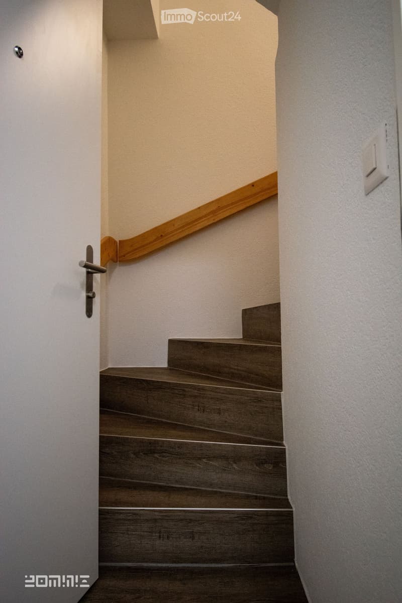 Wooden staircase with a handrail, leading upwards, with a door visible on the left side.