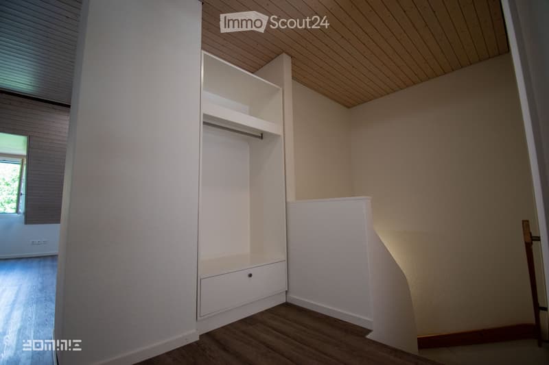 Empty room with wooden flooring, white built-in closet with shelves and drawers, and a slanted wooden ceiling.