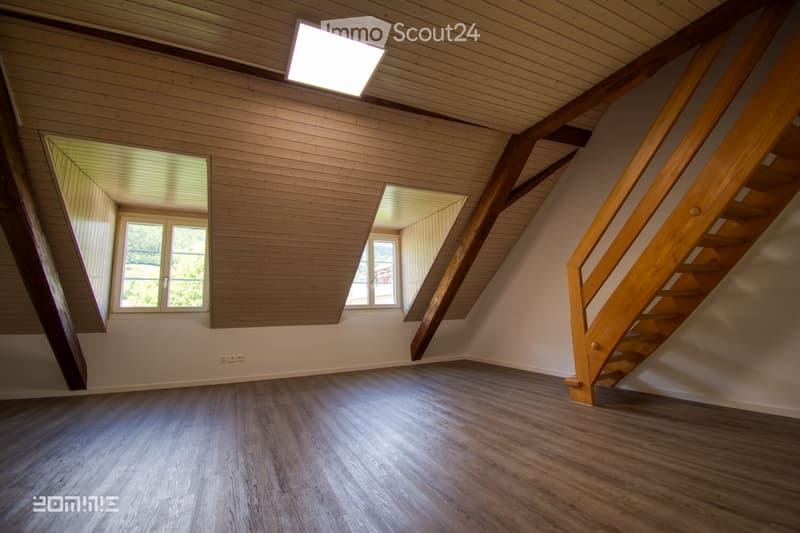 Empty attic space with wooden walls, slanted wooden ceiling, wooden staircase, and large windows letting in natural light.