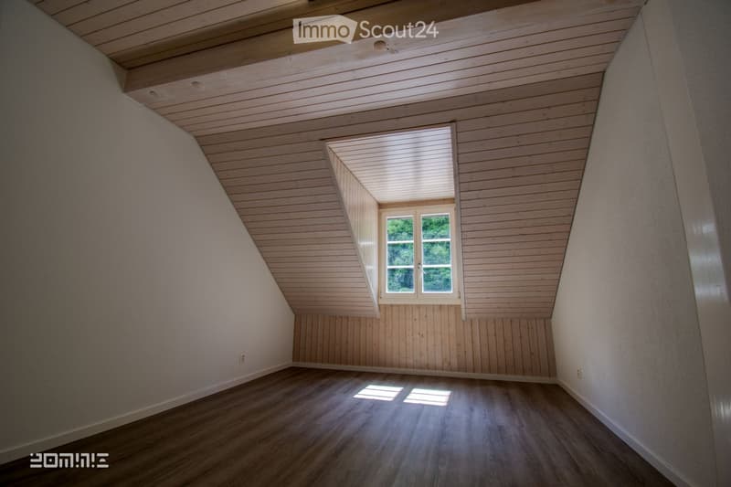 Empty attic room with wooden walls, ceiling, and floor, featuring a single window with white blinds and a view of greenery outside.