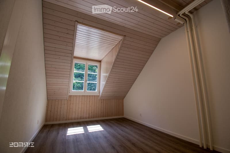 Empty attic room with wooden walls, slanted wooden ceiling, and a single window letting in natural light. The floor is wooden, and the walls are painted in neutral tones.