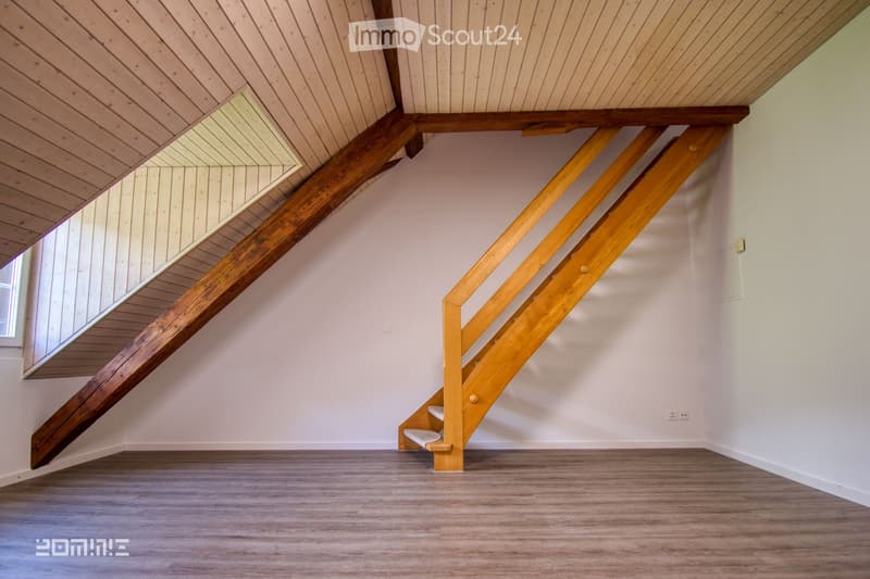 Empty room with wooden floor, white walls, and a wooden staircase leading to an attic space with exposed wooden beams and slanted ceiling.