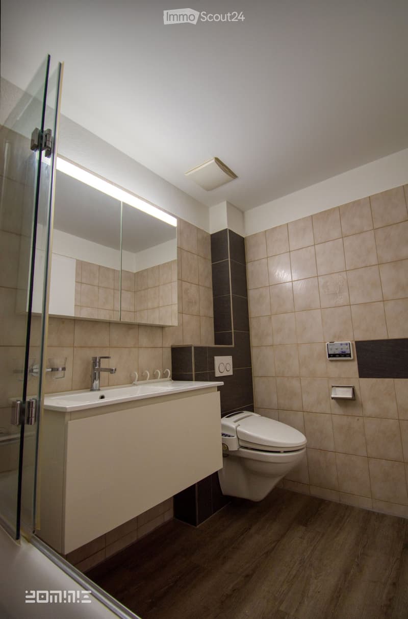 Modern bathroom with beige tiles, a double sink, a toilet, a large mirror, and a glass shower enclosure.