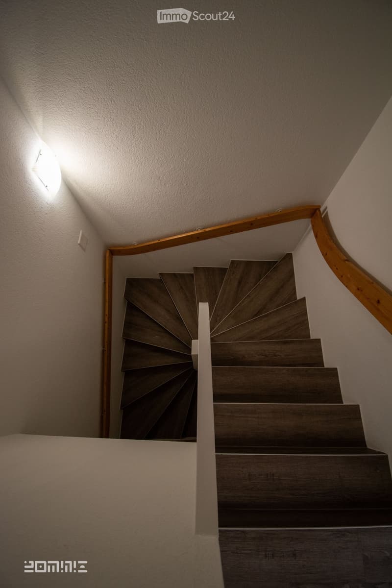 Spiral staircase with wooden steps and handrail, illuminated by a wall-mounted light fixture.