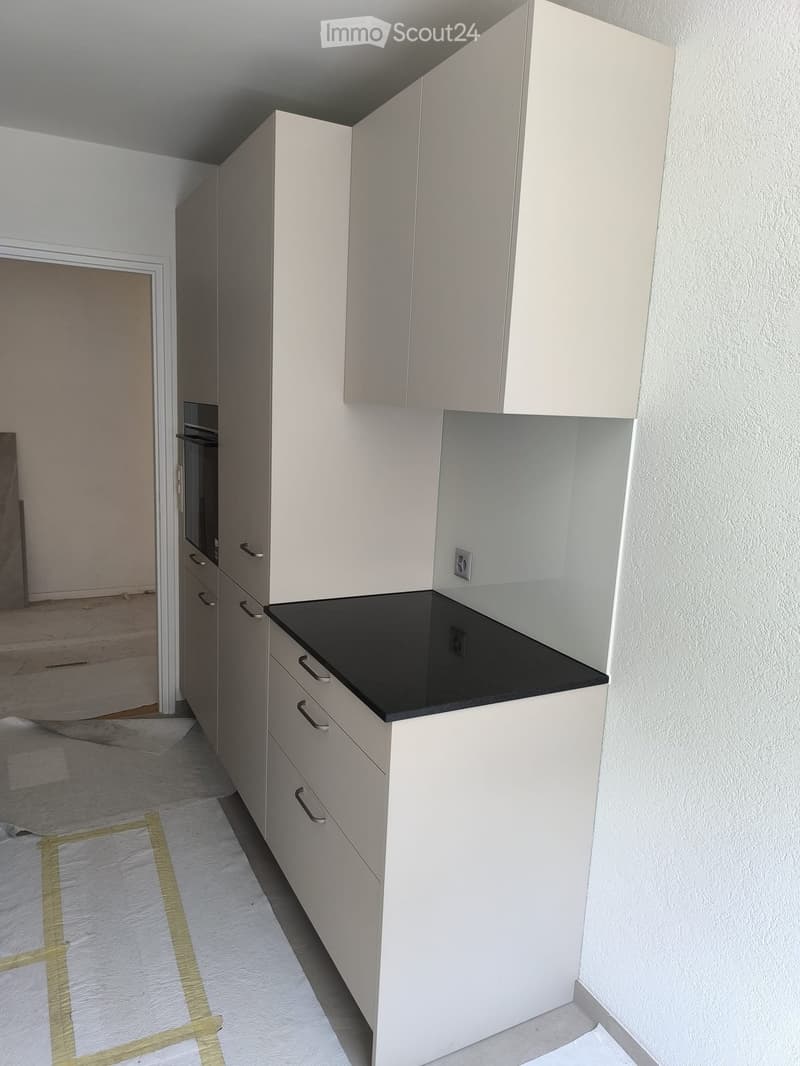 Empty kitchen area with white cabinets, black countertop, and a partially visible oven in the background. The floor is covered with protective material.