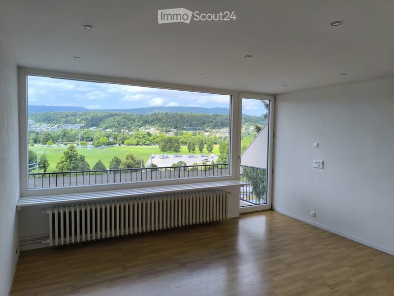 Empty room with wooden flooring, large window offering a scenic view, radiator, and balcony access.