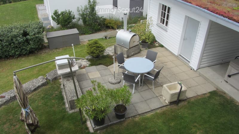 A well-maintained garden with a patio area featuring a round table, four chairs, a barbecue, and potted plants. The space includes a grassy lawn, a small shed, and a modern house with white siding.