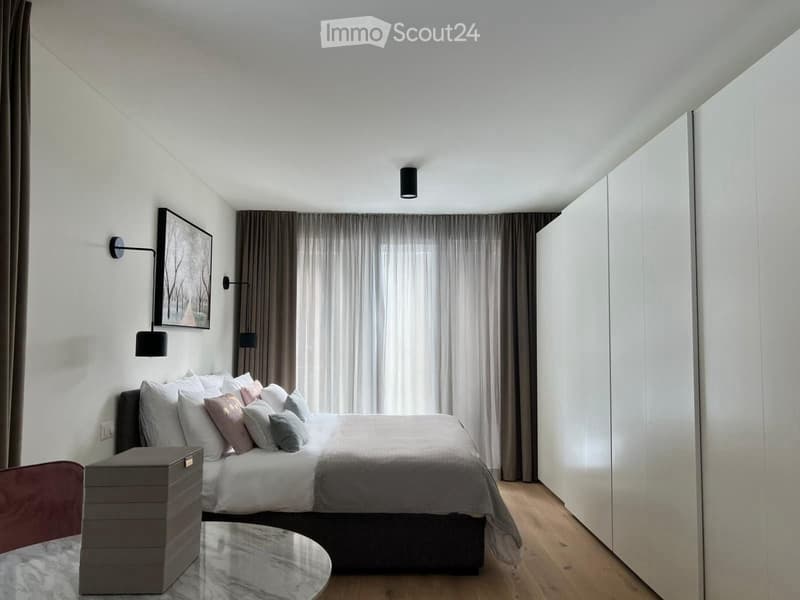 Bedroom with bed, white walls, wooden floor, large windows, wall-mounted lamps, picture frame, white and grey bedding, white wardrobes, round table with storage box