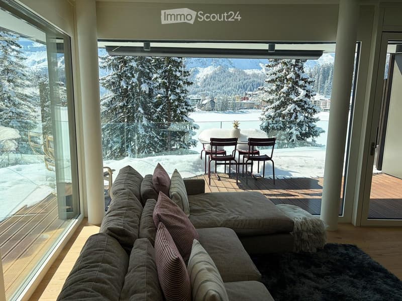 large living room with sofa, coffee table, chairs, table, glass walls, balcony, wooden floor, snowy mountains view