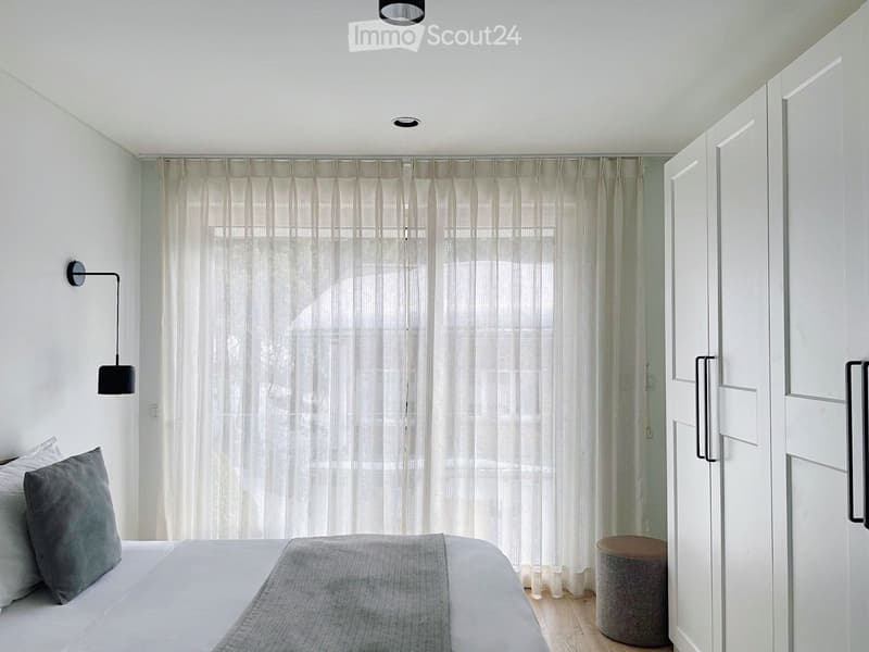 Bedroom with white walls, wooden floor, bed, white curtains, wall lamp, white and gray pillows, white wardrobe, ottoman