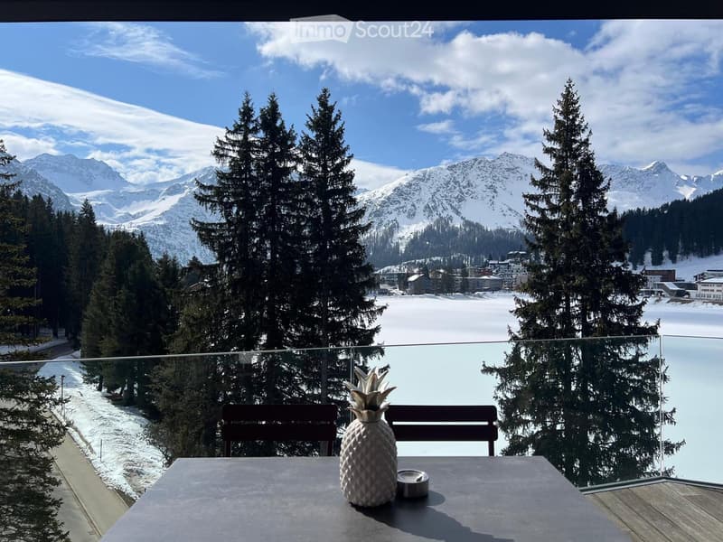 Outdoor dining area with a table, chairs, and a glass railing overlooking a snowy landscape with mountains and trees.