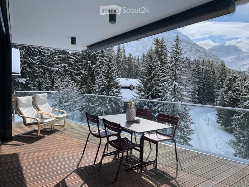 wooden deck, glass railing, table with 4 chairs, 2 lounge chairs, snow-covered mountains in the background