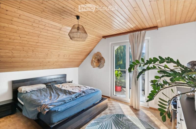Cozy attic bedroom with wooden slanted ceilings, a large bed with blue bedding, a nightstand, a wicker pendant light, a wall-mounted decorative piece, a potted plant, and a dresser.