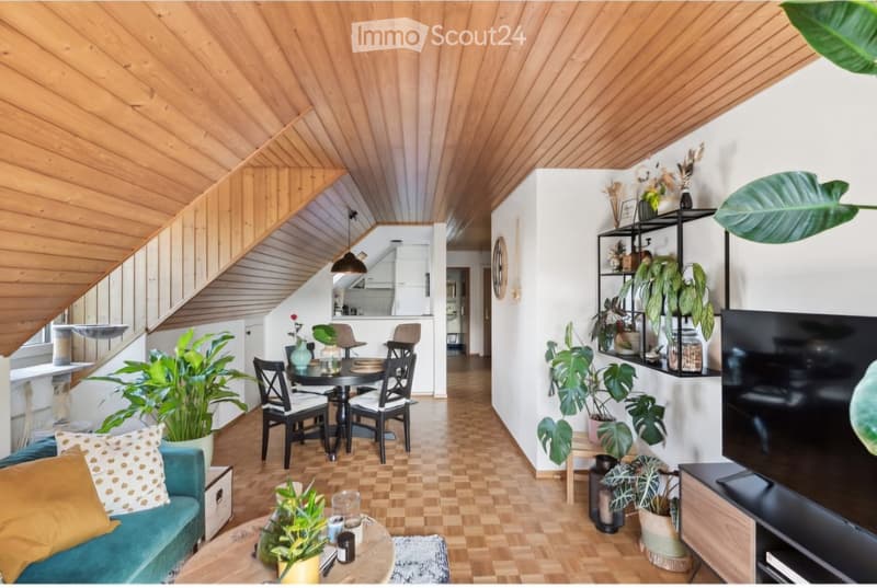 A modern, open-concept living and dining area with wooden flooring, a slanted wooden ceiling, and multiple potted plants. The living area features a teal sofa with patterned cushions, a round coffee table, and a television on a wooden stand. The dining ar