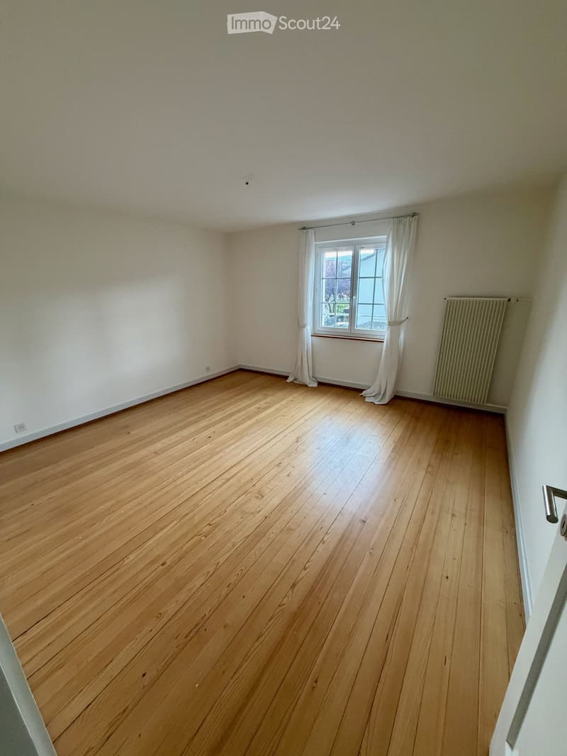 Empty room with wooden flooring, white walls, a window with white curtains, and a radiator on the wall.