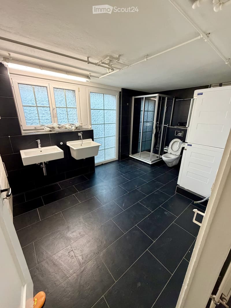 Modern bathroom with dark tiled floors and walls, featuring two sinks, a toilet, a glass-enclosed shower, and a window with frosted glass panels.