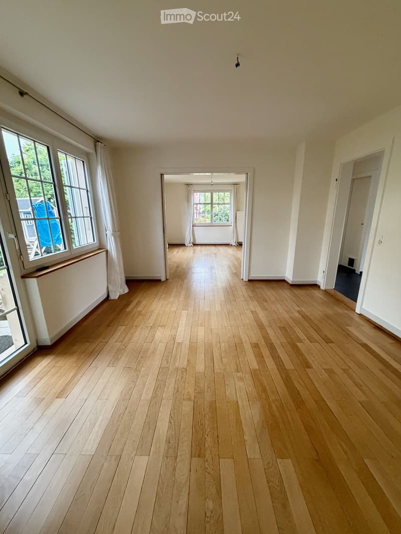 Empty hallway with light wooden flooring, white walls, large windows with white curtains, and a door leading to another room.