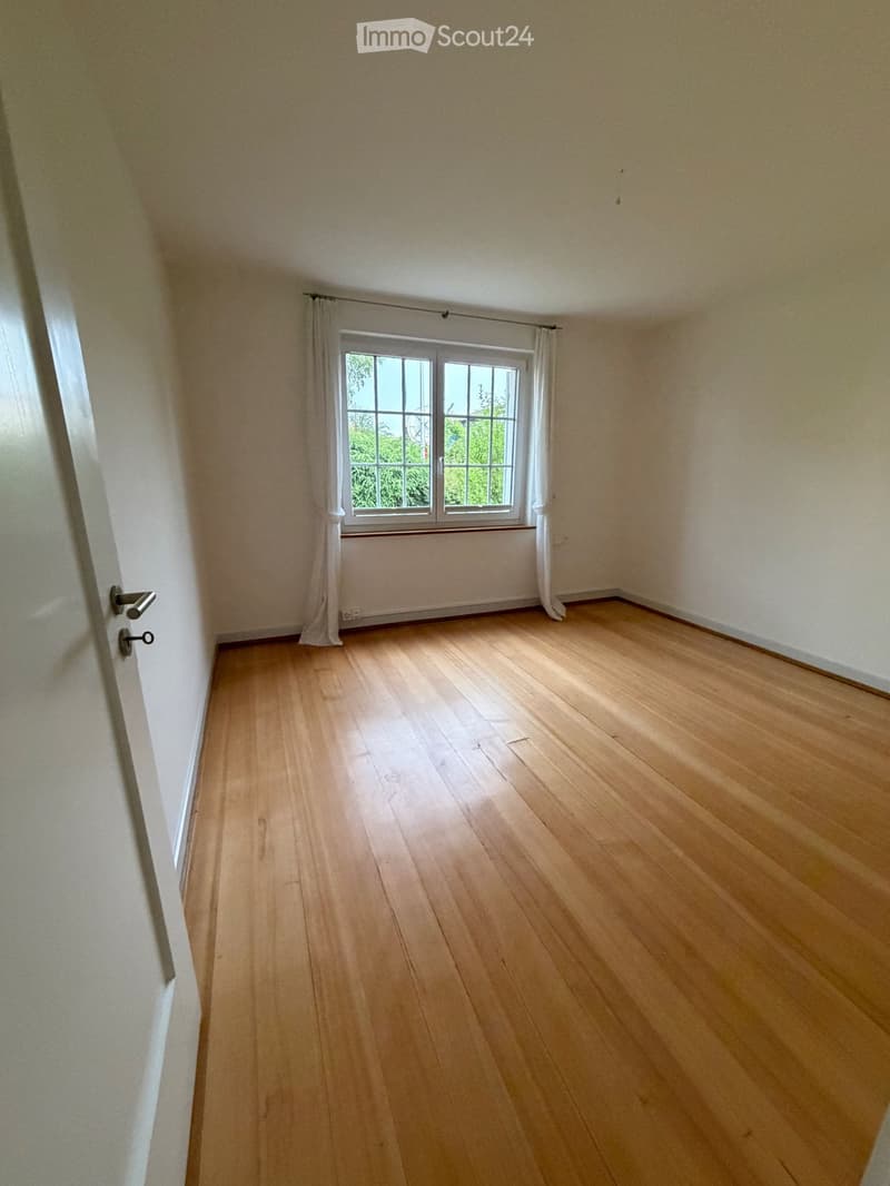 Empty room with wooden flooring, white walls, and a window with white curtains. The room has a door on the left side.