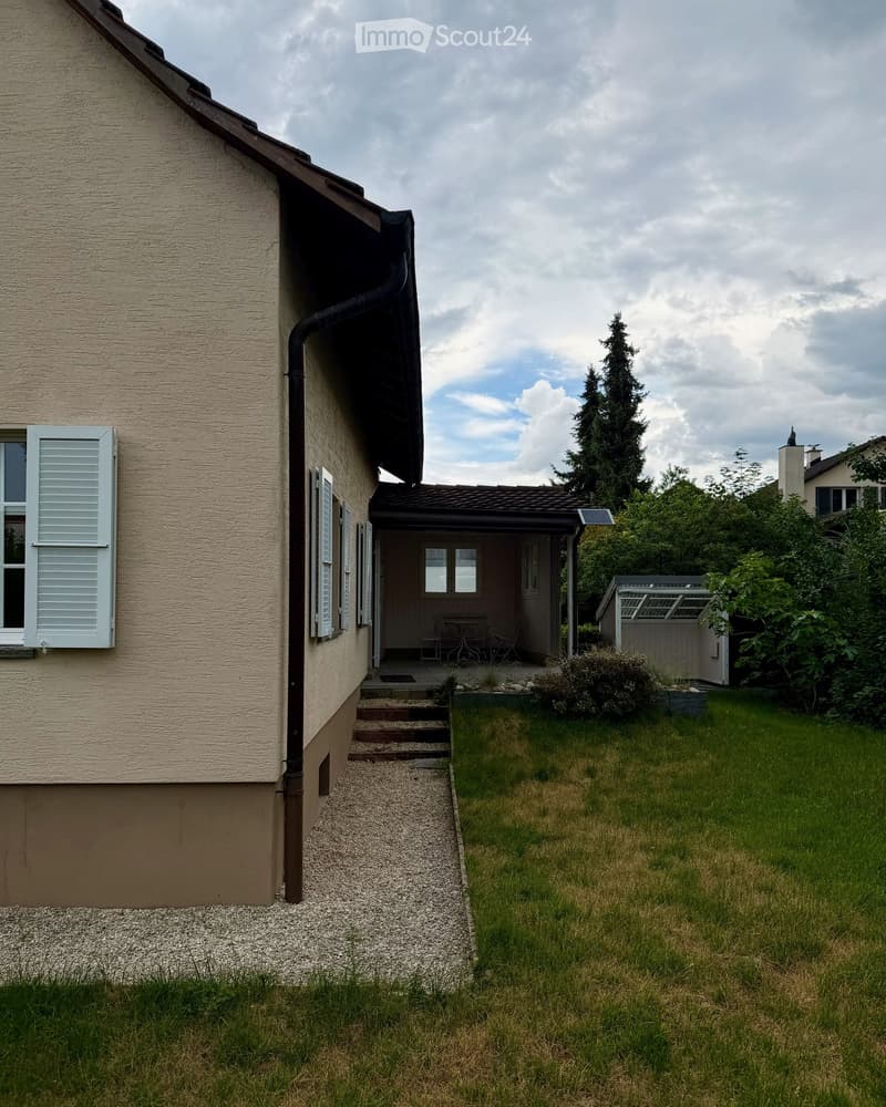 Side view of a house with a gravel pathway leading to a covered patio area, small garden, and surrounding greenery.