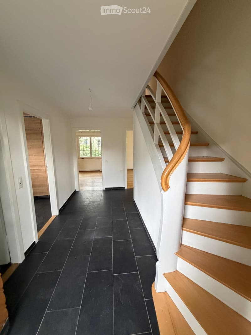 Empty hallway with black tiled floor, white walls, wooden staircase with handrail, and a window at the end.