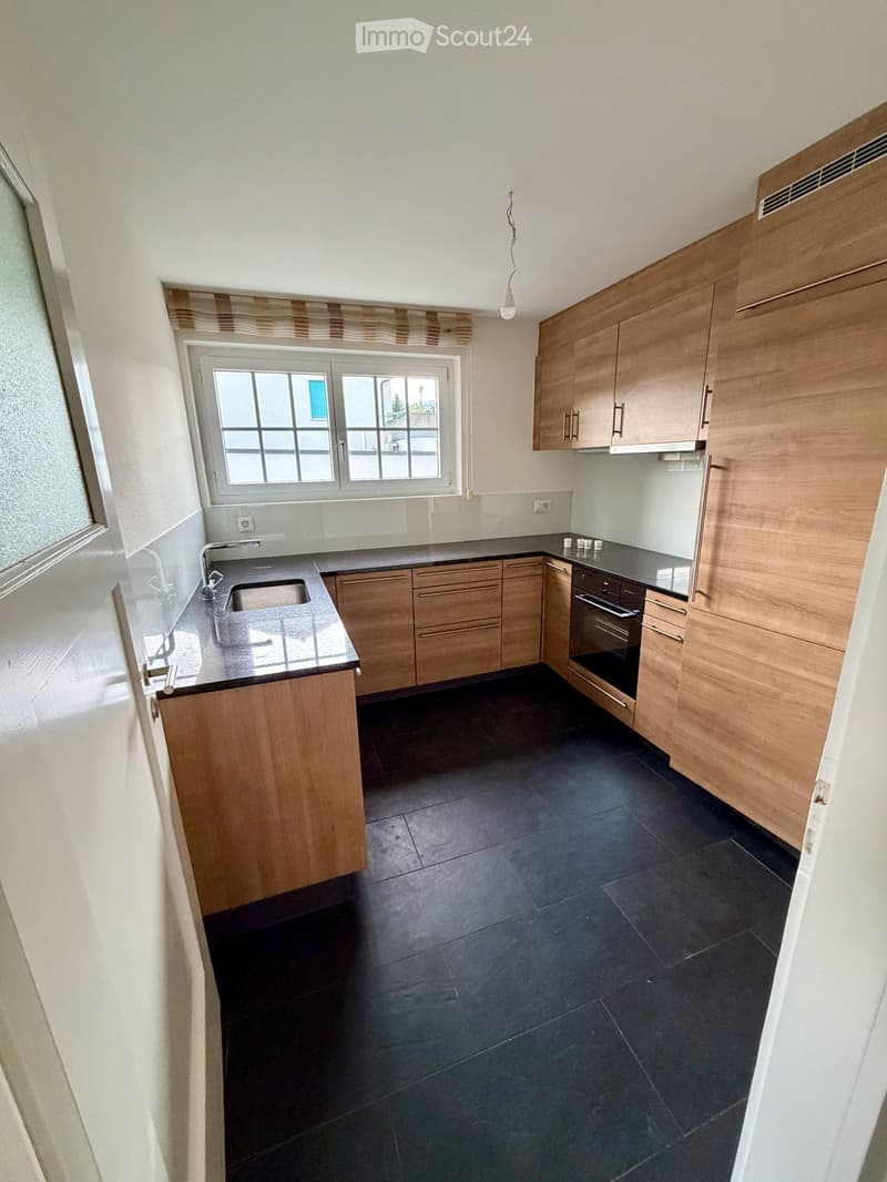 Modern kitchen with wooden cabinets, black countertops, and a stainless steel sink. The floor is dark tiled, and there is a window above the sink.