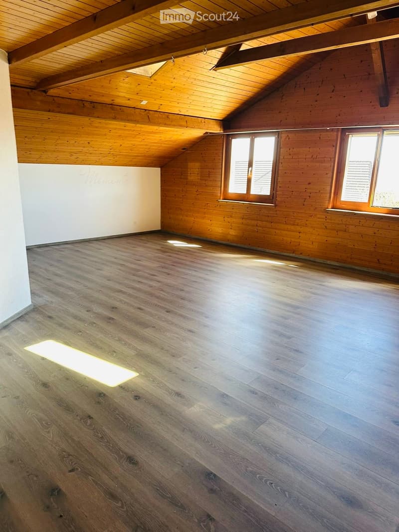 Empty room with wooden floors, exposed wooden beams on the ceiling, and large windows allowing natural light to enter.