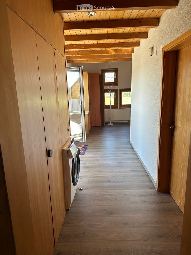 A long wooden hallway with exposed wooden beams on the ceiling, wooden flooring, and a private washing machine on the left side. The hallway has several doors and windows, with a small rug near the washing machine.