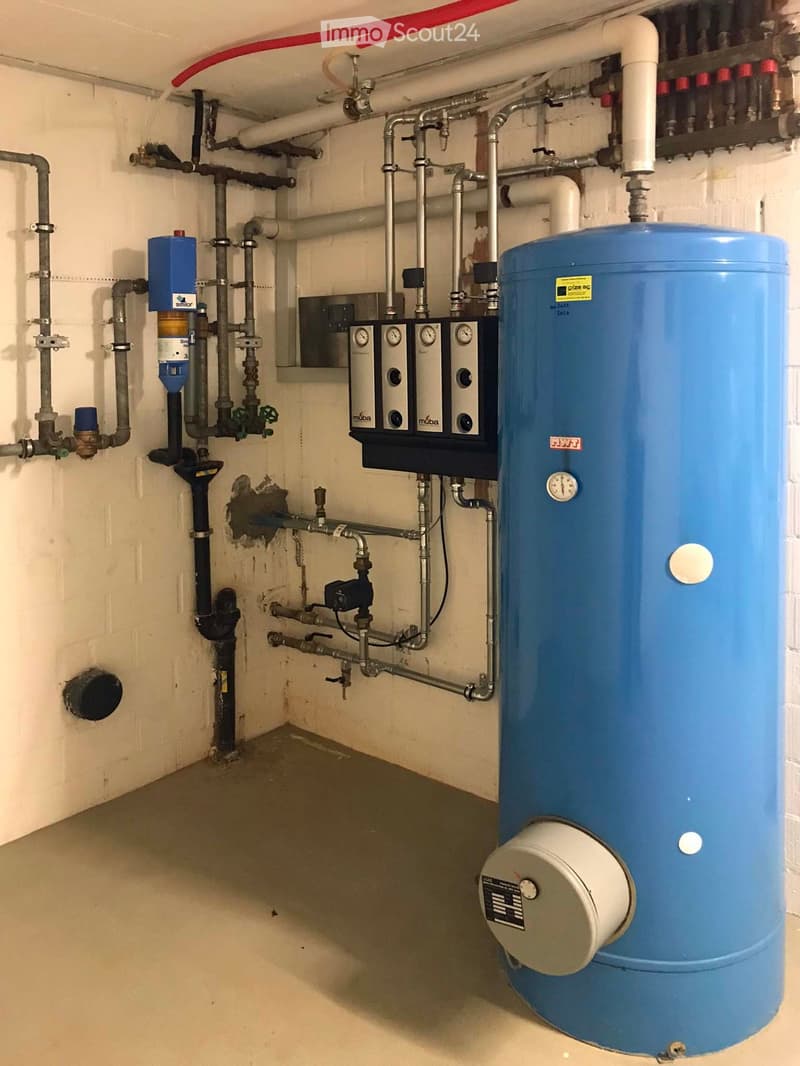 The image shows a utility room with various plumbing and heating equipment, including a large blue water heater tank, valves, and pipes. There is also a washing machine and what appears to be a dishwasher visible in the room.