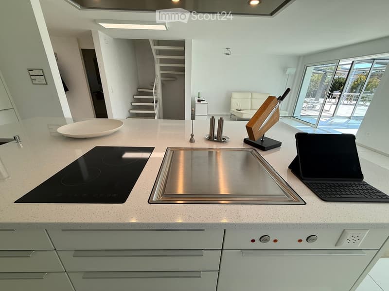 Modern kitchen with white countertops, stainless steel sink, induction cooktop, and built-in dishwasher. The kitchen also features a tablet or laptop on the counter, indicating a home office or work area.
