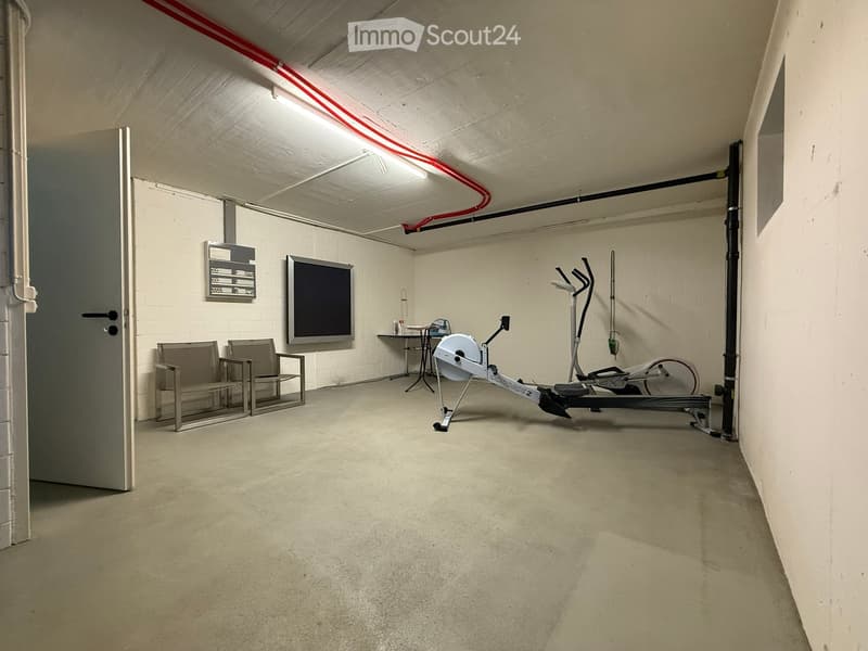 The image shows a garage or basement-like space with concrete floors, white walls, and various exercise equipment such as an elliptical machine and weight training equipment. There are also some chairs and a TV mounted on the wall, suggesting this space m