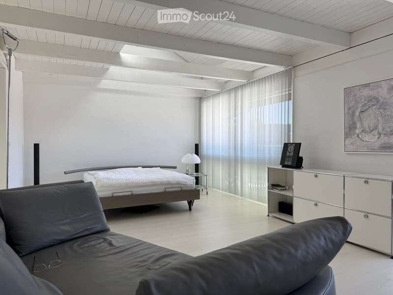 Spacious bedroom with white walls, wooden ceiling beams, large windows, grey leather sofa, modern bed frame, and built-in storage units