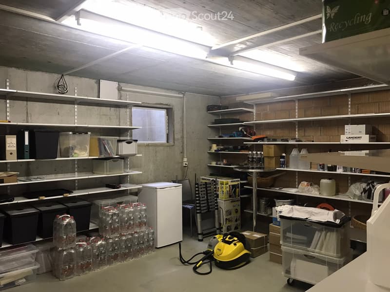 This image shows a storage room or basement area with various shelves and storage containers. The room has concrete walls and a ceiling with exposed beams and lighting. There are several plastic storage bins, a refrigerator, and a yellow cleaning machine