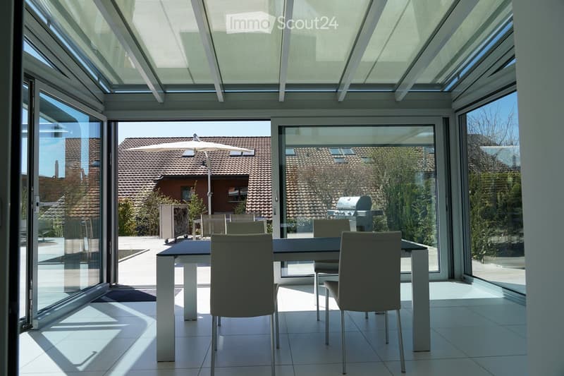 Modern glass-enclosed dining area with a view of the backyard, which includes a garage and an outdoor kitchen/grill. The room has a sleek, minimalist design with a large dining table and chairs.