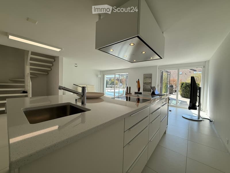Modern, open-concept kitchen with white cabinets, quartz countertops, stainless steel sink, and built-in dishwasher. The kitchen features recessed lighting and overlooks a patio or outdoor area through large sliding glass doors.