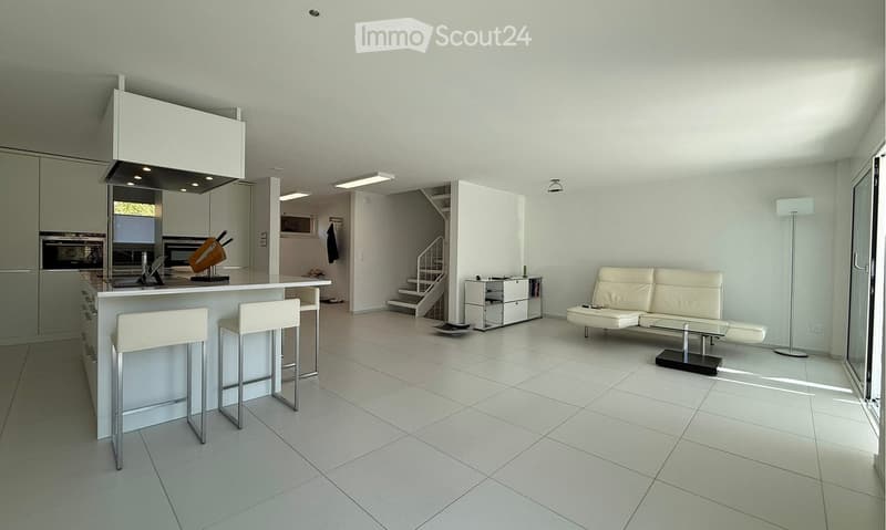 Spacious open-plan living area with modern kitchen, dining space, and living room. The kitchen features built-in appliances including a dishwasher. The living room has a white leather sofa and other furniture. The space has a minimalist, contemporary desi