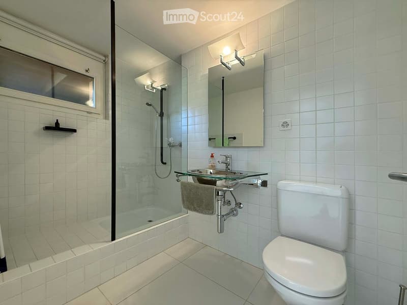 Spacious bathroom with white tile walls and floors, glass shower enclosure, vanity with glass countertop, and toilet. Lighting fixtures and mirror above the vanity.