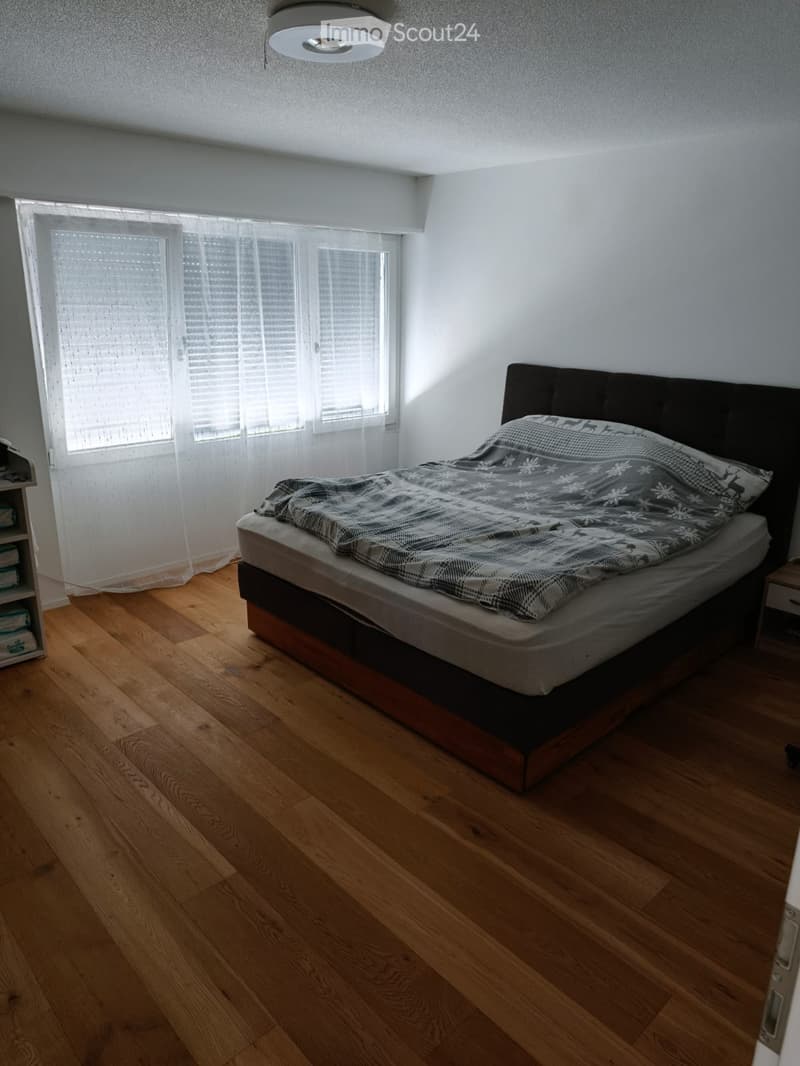 Bedroom with wooden flooring, large window with blinds, bed with dark headboard and patterned bedding, nightstand, and a shelf with folded towels.