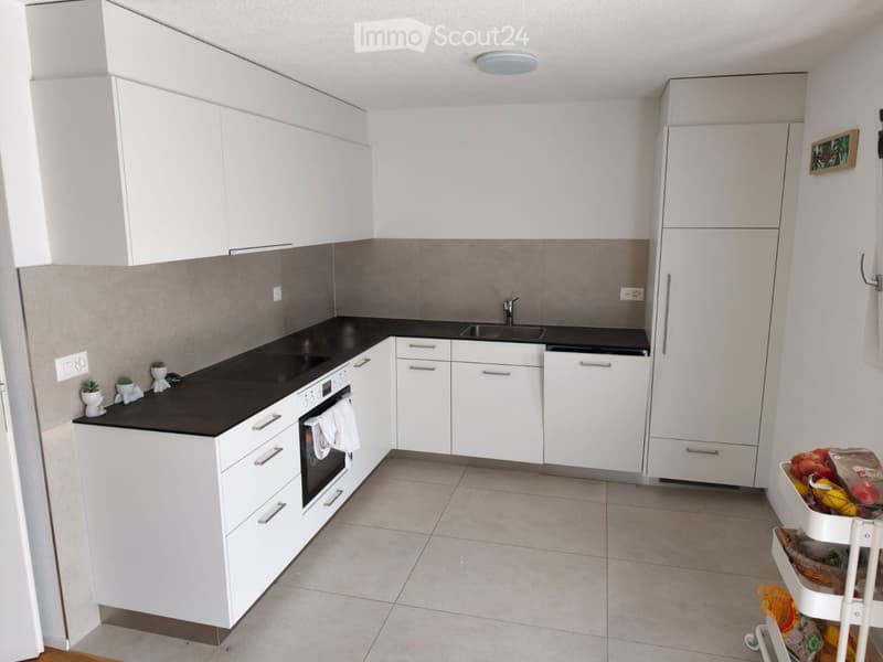 Modern kitchen with white cabinets, black countertops, and a built-in oven. A small cart with fruits and vegetables is visible in the corner.