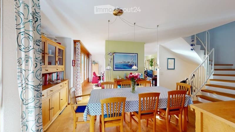 Bright dining room with wooden chairs around a table, a cabinet with glass doors, a picture frame on the wall, and a staircase leading upstairs.
