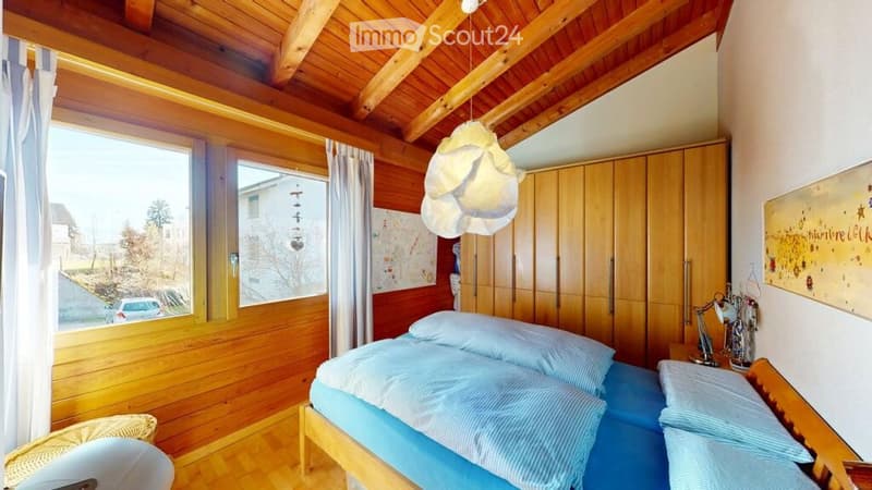 Cozy bedroom with wooden walls and ceiling, large window with curtains, bed with blue bedding, wardrobe, nightstand with lamp, and a wicker basket on the floor.