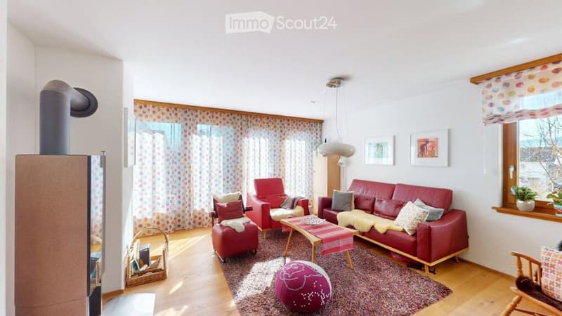 Bright living room with wooden flooring, red leather sofa, armchair, coffee table, ottoman, and a fireplace. Decor includes a colorful ball, picture frames, and curtains.