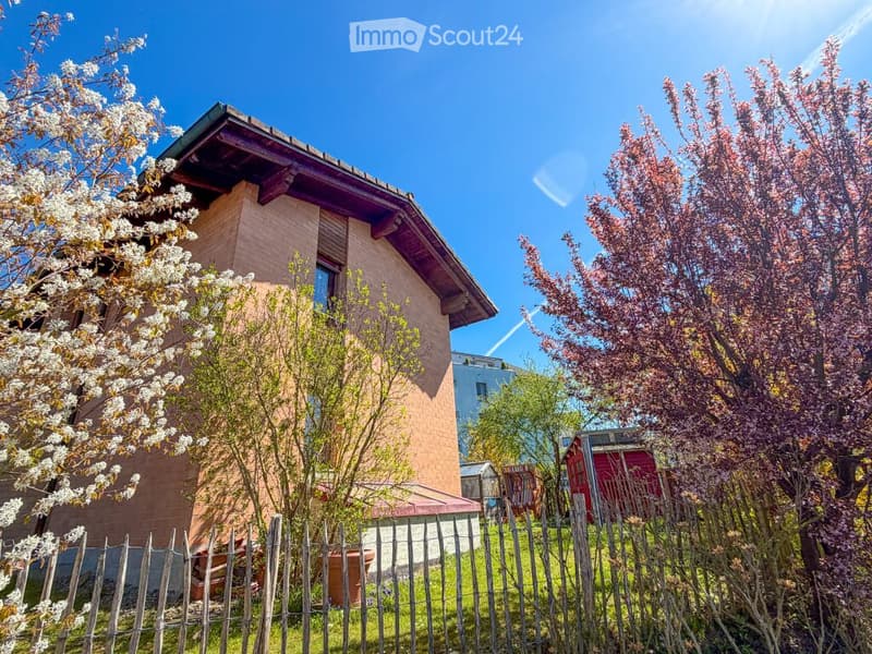 A two-story house with a brick facade and wooden roof, surrounded by a wooden fence, flowering trees, and shrubs.